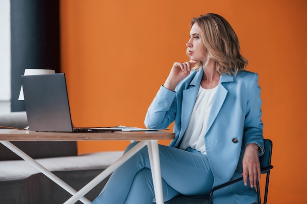 Businesswoman with curly blonde hair indoors in room with orange colored wall and wooden table.