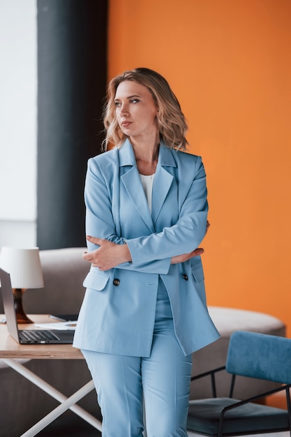 Businesswoman with curly blonde hair indoors in room with orange colored wall and wooden table.