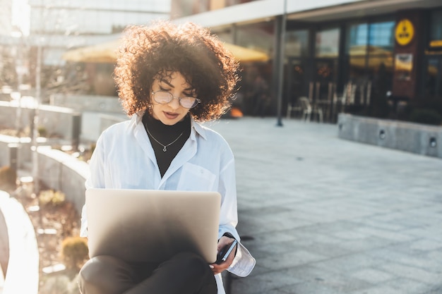 Businesswoman with curly hair and eyeglasses working outside with a laptop in a sunny day