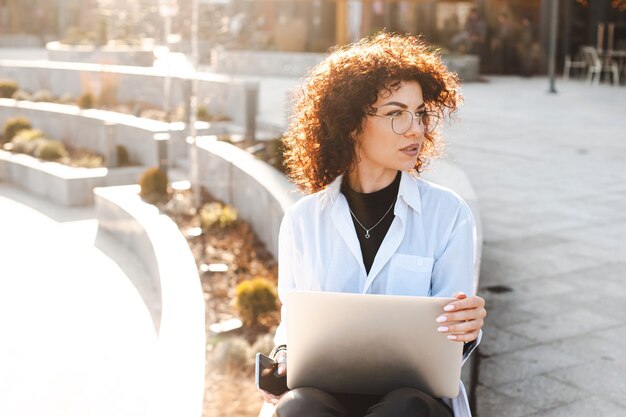 Businesswoman with curly hair working at laptop outside for fashion lifestyle design mobile communic...