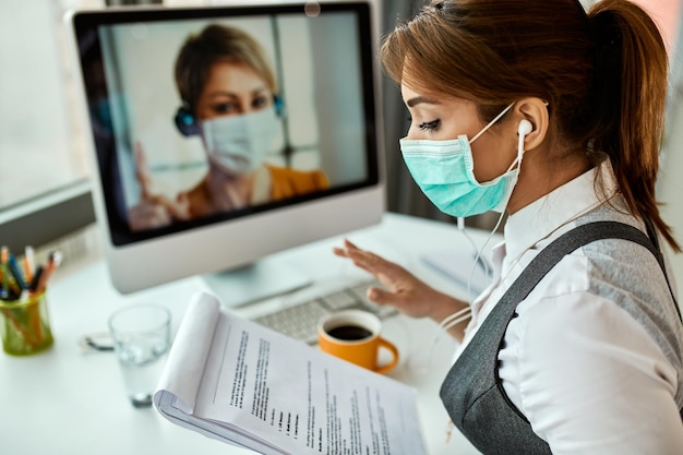 Businesswoman with face mask going through reports while having online meeting with her colleague in the office