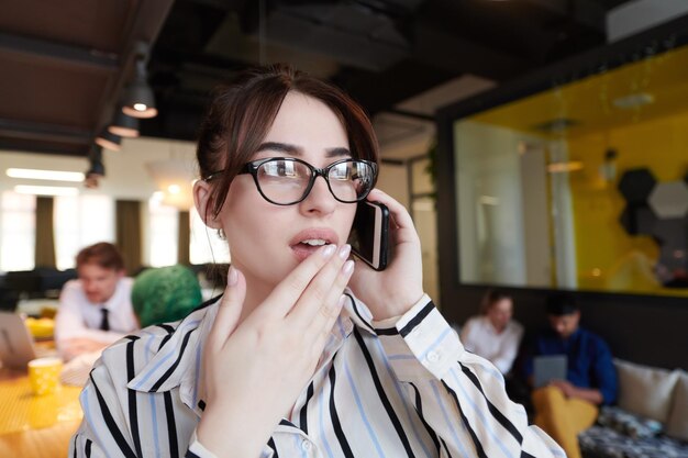 현대 스타트업 오픈 플랜 사무실 내부에서 휴대폰을 사용하는 안경을 쓴 여성 사업가