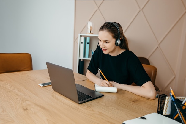 Businesswoman with headphones on sits at desk and looks at laptop taking notes at home