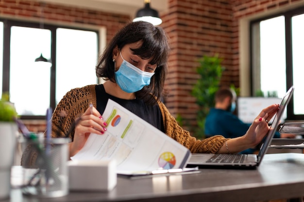 Деловая женщина с медицинской маской для предотвращения заражения коронавирусом смотрит на статистический документ компании, работающий над маркетинговой стратегией. предприниматель женщина анализирует бизнес-отчет