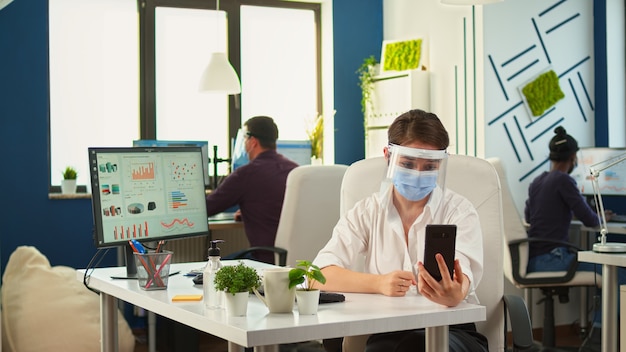 新しい通常のオフィスに座っているリモートの従業員と話しているビデオ会議のためのワイヤレスヘッドフォン付きのスマートフォンを使用して保護マスクを持つ実業家。社会的距離を尊重して働く同僚