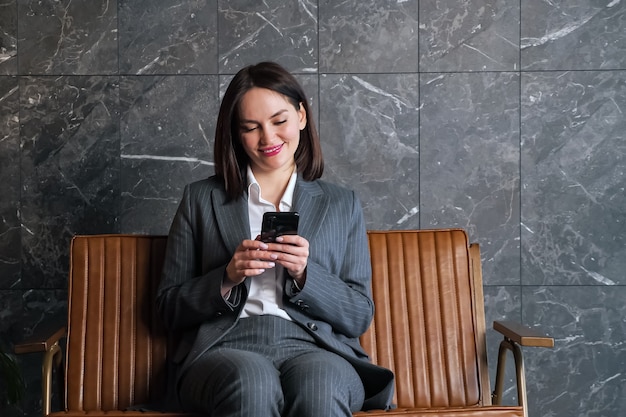 Деловая женщина с короткими темными волосами на черном смартфоне сидит на коричневой скамейке на фоне серо-белой мраморной стены в приемной нового офиса