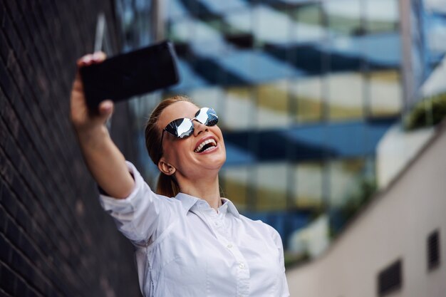 Businesswoman with sunglasses standing outside and taking selfie. Business center exterior.