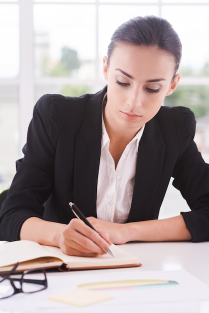 Imprenditrice al lavoro. fiduciosa giovane impresa che scrive qualcosa su carta mentre è seduta al suo posto di lavoro