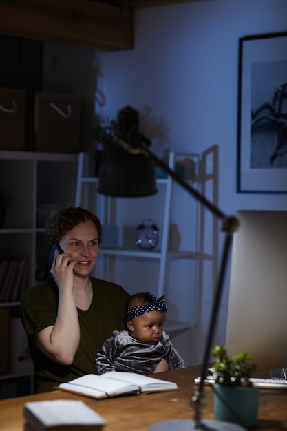 Деловая женщина, работающая дома до ночи, разговаривает по мобильному телефону, сидя за столом со своей маленькой дочерью