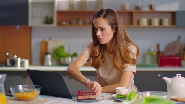 Businesswoman working on laptop computer at home office Portrait of thoughtful girl eating cake on domestic kitchen Pretty business woman using laptop on kitchen in slow motion