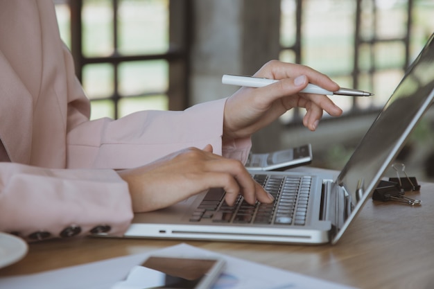 Businesswoman working on laptop in her workstation.Black blank screen smartphone, potted plant, pencil, notes, earphone on wooden desk. Close up