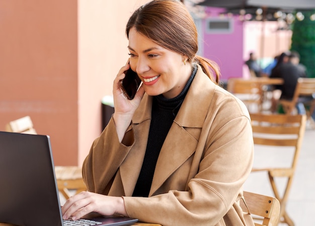 Photo businesswoman works on laptop in outdoor workspace speaks on phone smiles wears brown coat