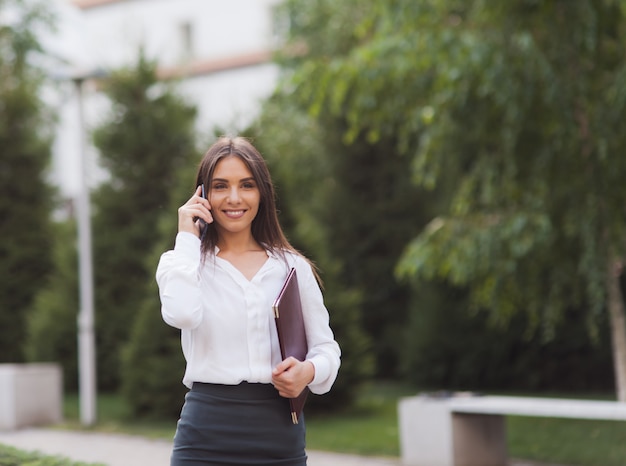 Businesswoman. Young female manager wearing a skirt and a blouse talking on phone at background of the city landscape. All in business