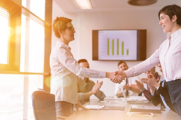 businesswomans handshake on team meeting  with group of people blured in background at modern startup business office interior