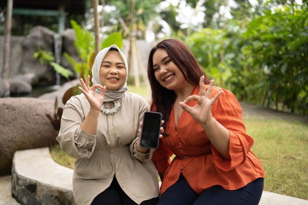 Photo businesswomen holding blank screen phone with okay hand gesture in outdoor cafe with happy