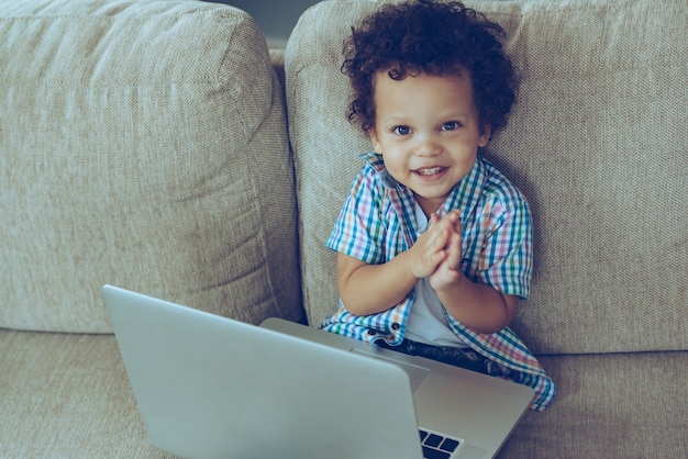 Beccato con il portatile! veduta dall'alto del piccolo bambino africano che guarda la telecamera con un sorriso mentre è seduto sul divano di casa con il computer portatile sulle ginocchia