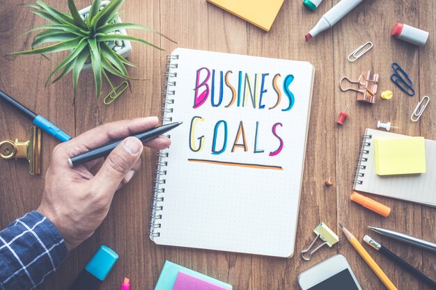 BUSUNESS GOALS message with male hand writing on notepad paper on wooden table and office supplies.Business concepts.flat lay design