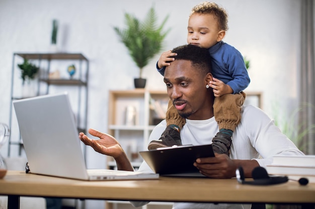 Photo busy afro man working on laptop with son on neck