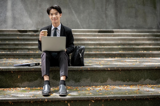 A busy Asian businessman sitting on stairs in front of the building working remotely in the city