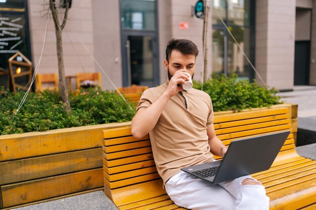 Busy bearded freelancer male remote working on laptop computer sitting on bench on city street and drinking takeaway coffee