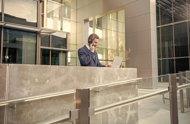 Занятый деловой человек в формальной одежде работает онлайн на компьютере, используя телефон для делового общения на открытом воздухе