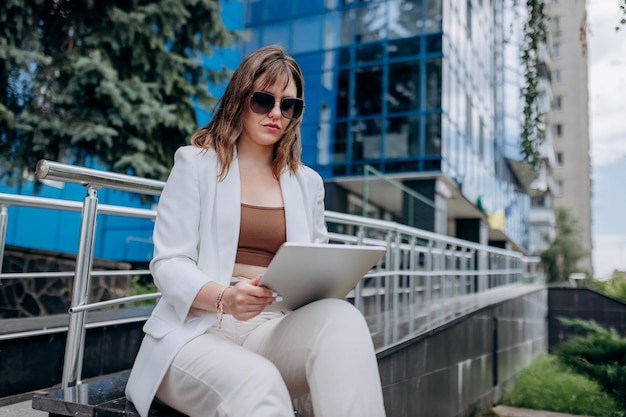 현대 사무실 건물 근처에 앉아 디지털 태블릿 작업을 하는 흰색 정장과 선글라스를 착용한 바쁜 비즈니스 여성