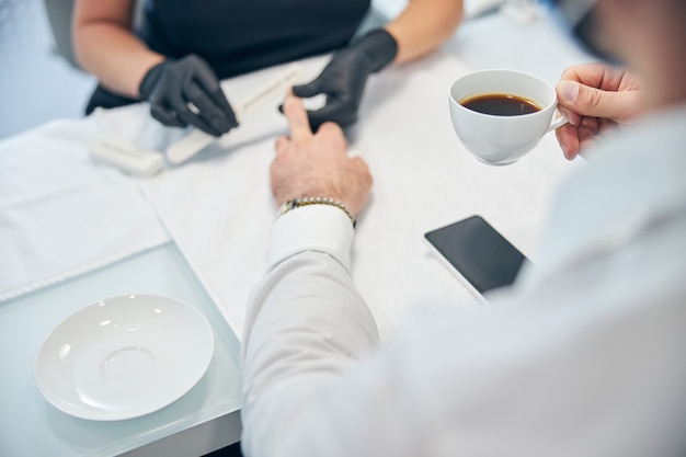 Busy businessman drinking coffee while being in salon on sterile manicure