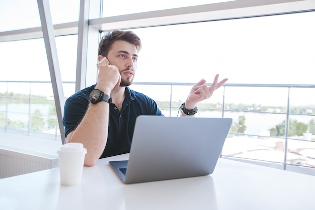 Busy businessman sits at the table near the window with a glass of coffee and a laptop, and talks on the phone.