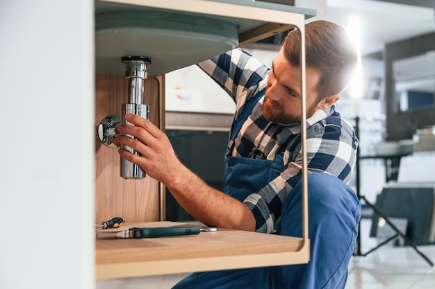 Photo busy by repairing siphon plumber in blue uniform is at work in the bathroom