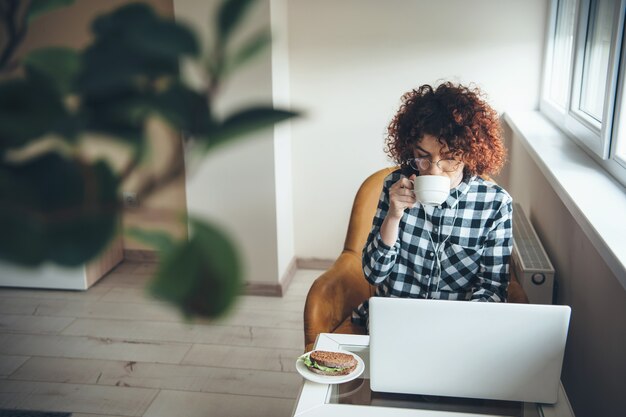 写真 ノートパソコンで作業中にサンドイッチとお茶を飲む巻き毛と眼鏡で忙しい白人女性