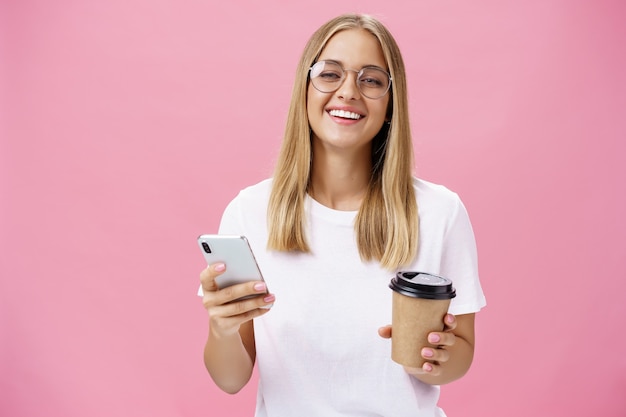 busy freelancer sipping coffee and working via smartphone talking to clients while sitting in cafe or coworking place, laughing joyfully at camera with broad smile holding paper cup and cellphone