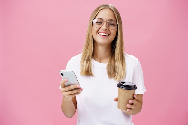 Busy freelancer sipping coffee and working via smartphone talking to clients while sitting in cafe or coworking place, laughing joyfully at camera with broad smile holding paper cup and cellphone