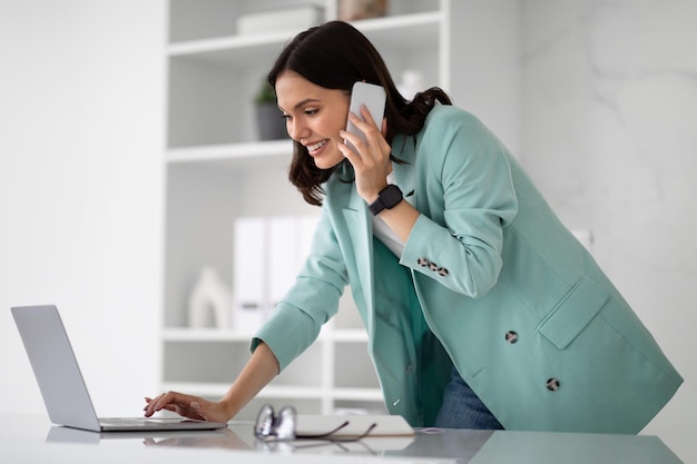 Busy happy young european woman in suit typing on laptop calling by phone