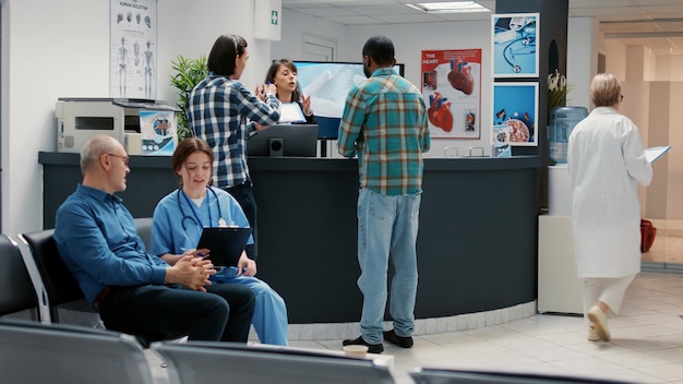 Busy hospital reception desk with diverse people in waiting\
room, asian and african american man using reports to attend\
medical appointment. waiting room lobby at facility clinic.