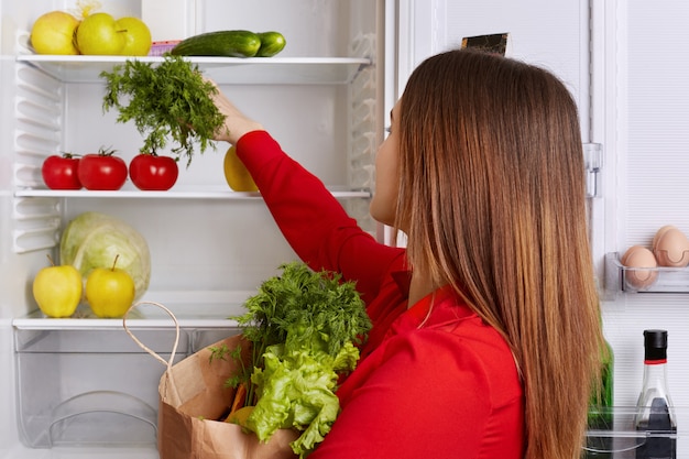 Busy housewife puts fresh vegetables she just bought on shelf of refrigerator