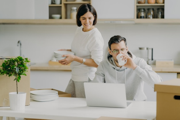 바쁜 남편은 노트북 컴퓨터 작업을 하고 커피는 안경을 쓰고 캐주얼한 스웨트셔츠를 입고 바쁘게 짐을 풀고 있는 아내는 이사하는 날 부엌에서 설거지를 하고 있다 가족과 모기지