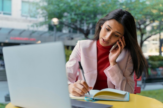 Busy Indian freelancer young serious businesswoman talking on mobile phone using laptop taking notes