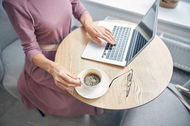 Busy lady sitting in front of her computer while typing message and going to drink coffee