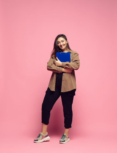 Busy and looks happy. Young woman in casual wear on pink background. Bodypositive character, feminism, loving herself, beauty concept. Plus size businesswoman, beautiful girl. Inclusion, diversity.