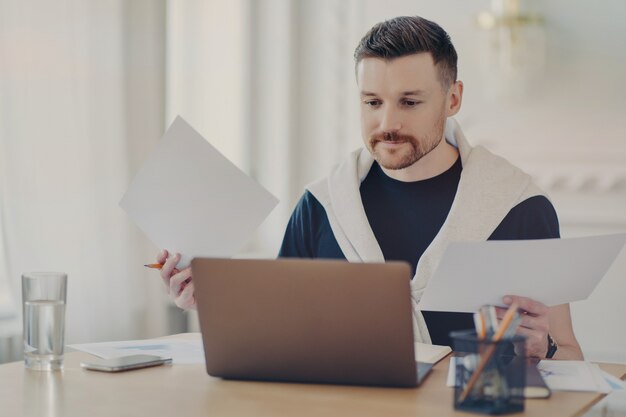 Libero professionista maschio impegnato in abiti casual seduto al suo posto di lavoro con il computer e facendo scartoffie, analizzando i risultati del progetto mentre lavora in remoto da casa. freelance e concetto di business