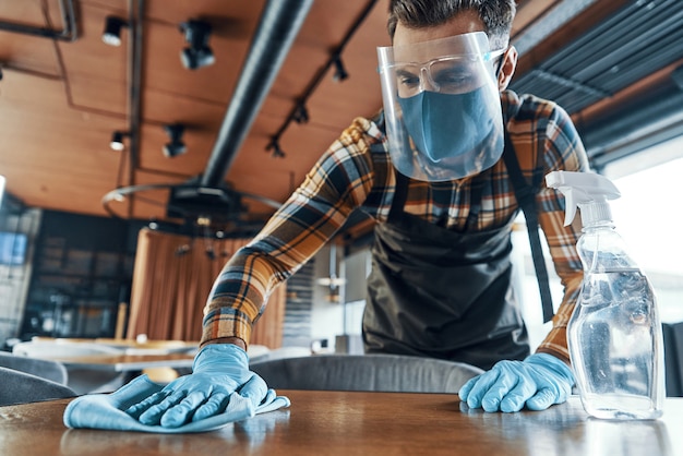 Busy man in protective face shield cleaning table in restaurant