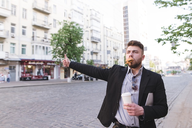 A busy man with a cup of coffee in his hands and a sandwich in his teeth catches a taxi on the road.