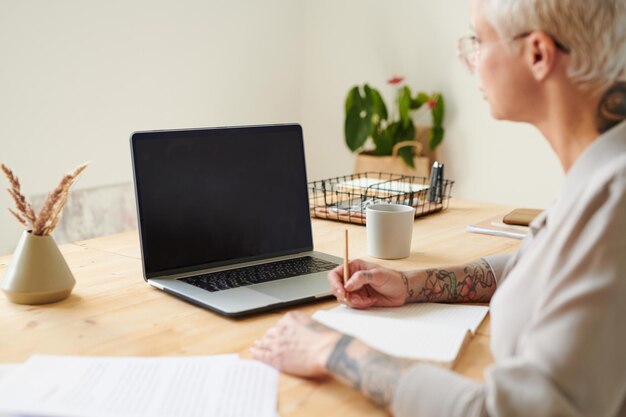 Busy middleaged woman sitting at desk and making notes in diary while studying to promote business w