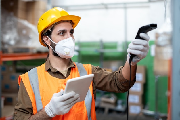 Busy mixed race warehouse employee in facial mask and gloves using digital tablet while scanning code of item