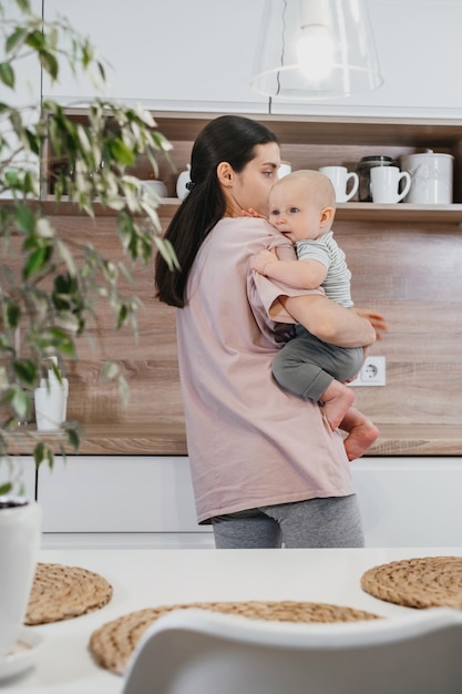 Busy mother cleaning kitchen with baby on her arms. Woman cooking and holding infant, tired woman doing daily homework with kid.