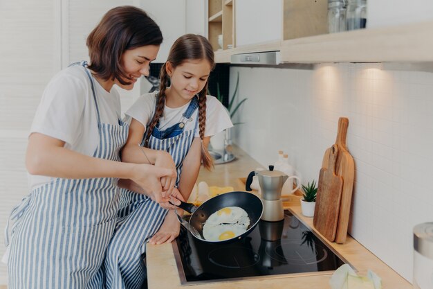 Busy mother and daughter wear striped aprons, pose at kitchen near cooker, fry eggs on pan, prepare fast breakfast