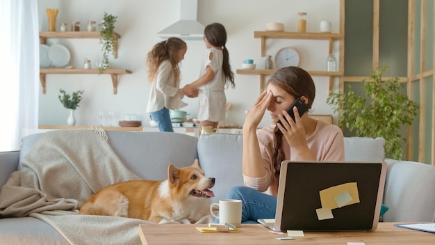 A Busy Mother Trying to Work Remotely With Her Children's at Home. Working From Home During Quarantine Epidemic. On The Background Her Daughters are Jumping Together Near Lying on the Couch a Cute Dog