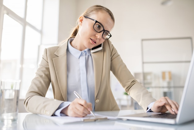 Busy multitasking young lady manager talking on phone, using laptop and making notes in paper in office, hardworking concept