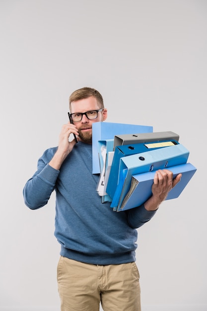 Busy office manager in casualwear holding stack of folders while consulting clients on smartphone in front of camera