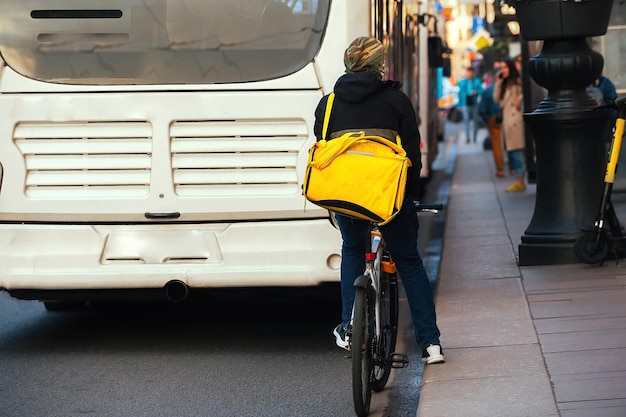 a busy part of a big city, a delivery man on a bicycle rides behind a bus with a yellow backpack